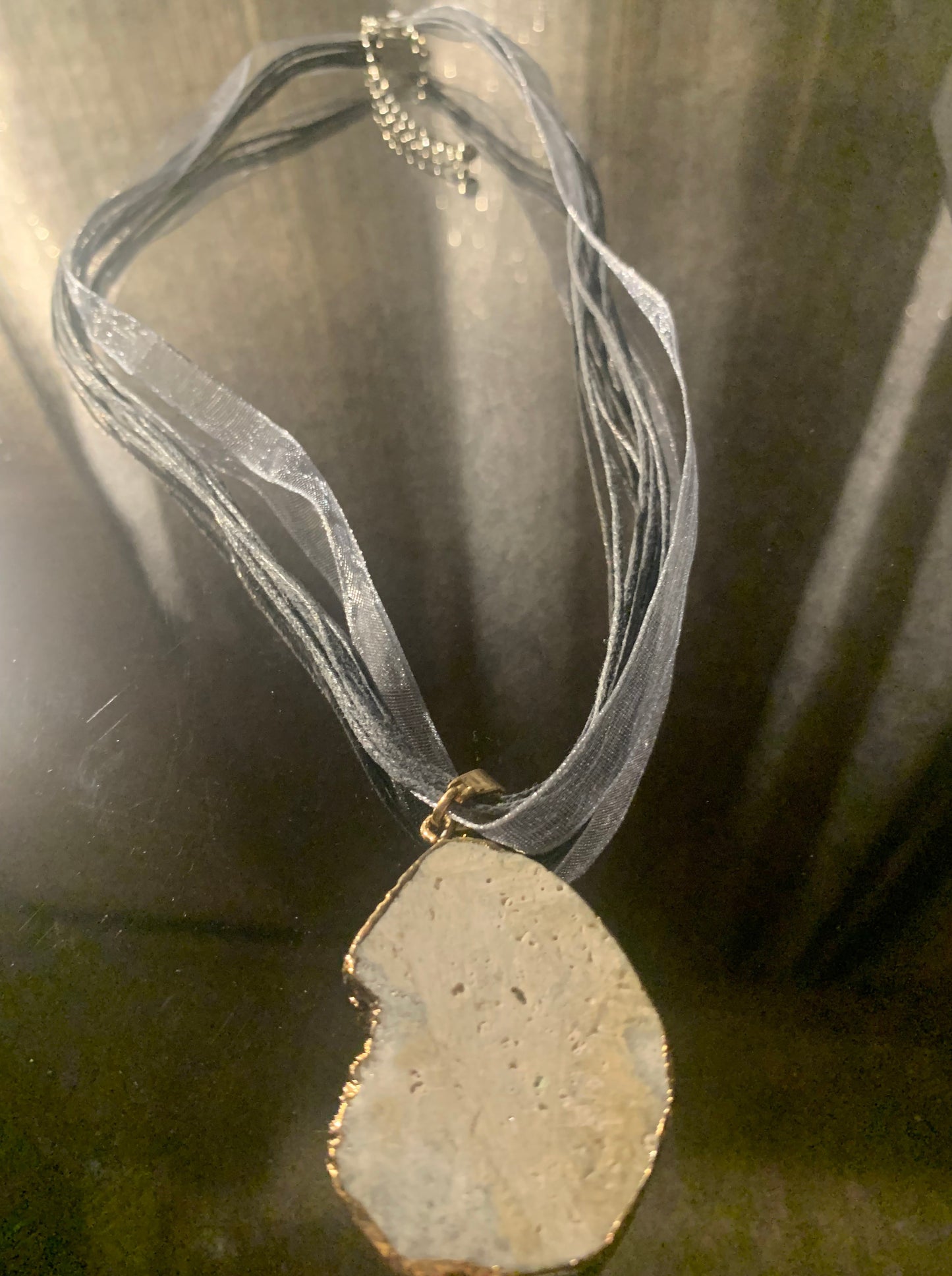 Necklace, Natural Pewter Cratered Stone framed in gold accented with 18" silver shimmer nylon ribbon multi cord finished with lobster clasp and extension chain.